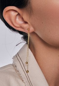 Złote wiszące kolczyki z dwoma kulistymi akcentami przymocowanymi do cienkich łańcuszków, prezentowane na delikatnie oświetlonym tle.