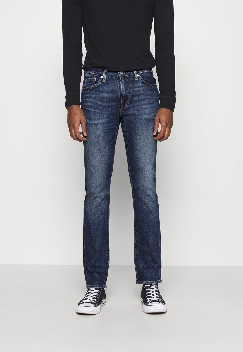Personne portant un jean slim bleu foncé, un haut noir à manches longues et des baskets noires et blanches, debout devant un fond uni.