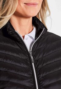 Schwarze Steppjacke mit gestepptem Design, hohem Kragen und silbernem Reißverschluss. Über einem weißen Hemd mit Kragen getragen. Glattes, leichtes Material.