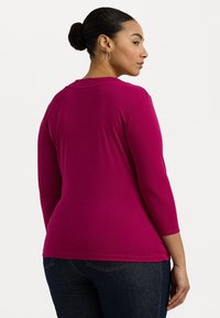 Fuksia langermet topp med rund hals og myk, glatt stoff. Har en figursydd silhuett og faller til hoften, i kombinasjon med mørk jeans.