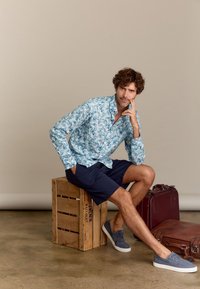 Wit overhemd met blauw bladdessin, marineblauwe shorts en grijze canvas schoenen. Zittend op een houten krat met een kastanjebruine koffer in de buurt.
