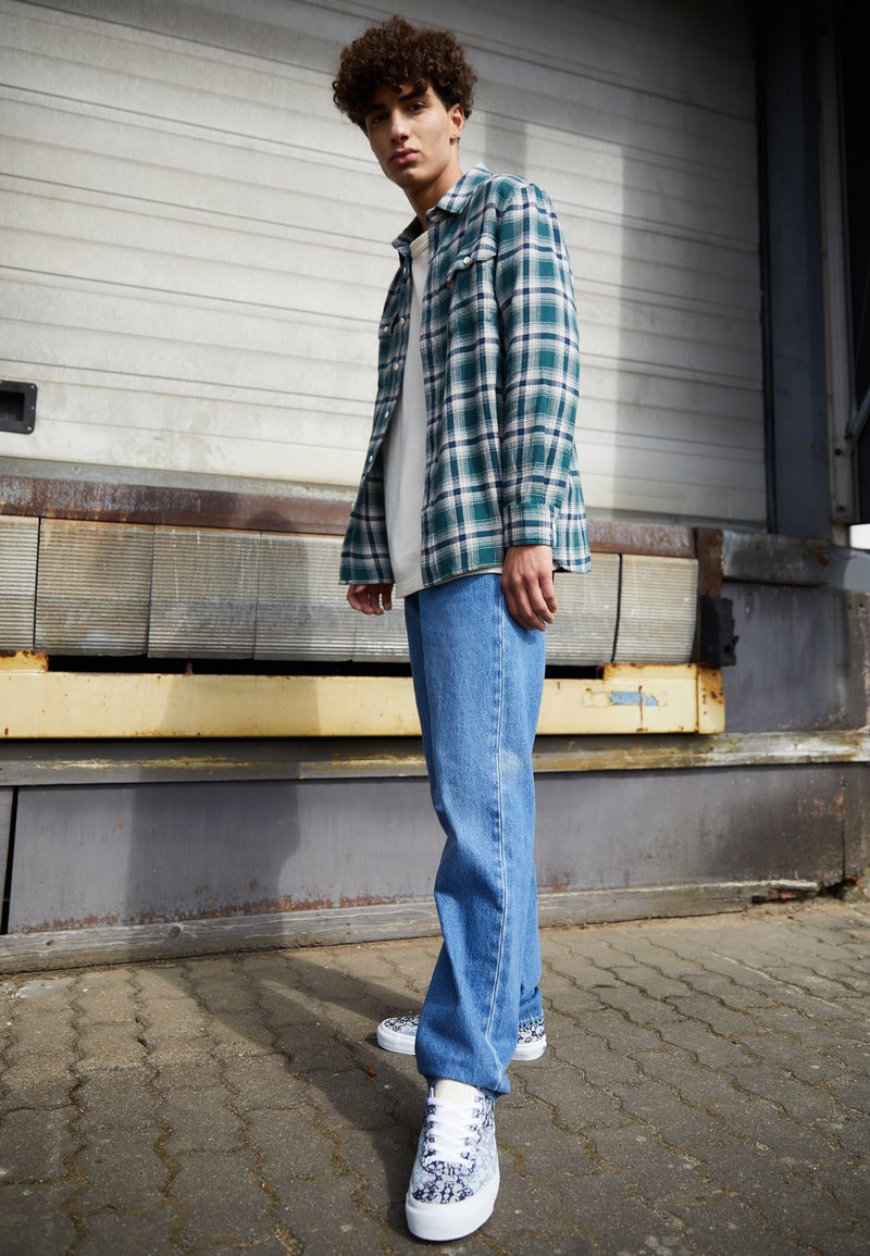 Groene en witte geruite flanellen shirt over een wit T-shirt, blauwe recht gesneden jeans en gemusterde sneakers tegen een gestructureerde achtergrond.