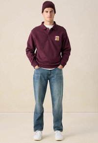 Bordeauxrot sweatshirt mit Kragen und Logopatch, kombiniert mit hellblauen Jeans. Eine Strickmütze und weiße Sneakers runden denLook ab.