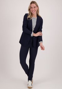 Marineblazer over een gestreept lange mouwen top, gecombineerd met marineblauwe broek en witte sneakers. Soepele stof met een afgestemde pasvorm en een sluiting met twee knopen.