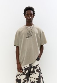 Béžové oversized tričko s grafickým potiskem listů a slovem "Hokusai." V kombinaci s volnými kalhotami s kravským vzorem.