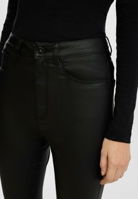 Pantalons en simili cuir noir avec taille haute, coupe slim, poches avant et passants de ceinture ; texture lisse et finition brillante.