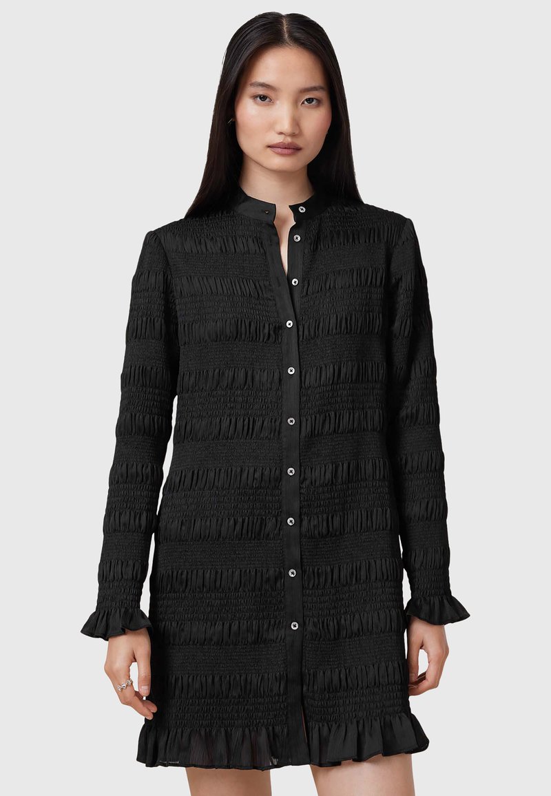 Abito camicia nero con texture a pieghe verticali, colletto alto, chiusura con bottoni e polsini arricciati. Presenta una vestibilità dritta e orlo a ginocchio.
