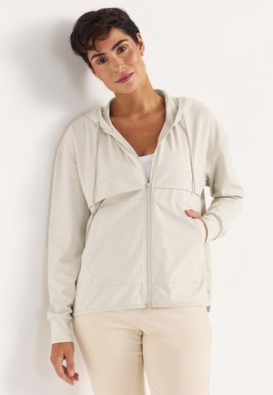 Frau mit kurzen dunklen Haaren, die einen hellbeigen Kapuzenpullover mit Reißverschluss und Taschen trägt, vor einem schlichten weißen Hintergrund stehend.