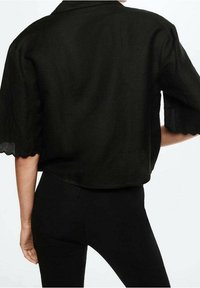 Schwarz geschnittenes Shirt aus strukturiertem Stoff, mit eingeschlitztem Kragen und gewellten Ärmeln. Getragen mit schwarzen, figurbetonten Hosen.