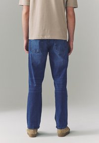 Blaue Jeans aus Denim mit gerade geschnittenen Beinen, ausgestattet mit Gesäßtaschen mit einem dezenten Y-Steppdesign, kombiniert mit einem hellbeigen T-Shirt.