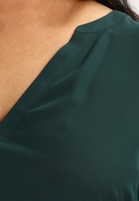 Blouse vert foncé en tissu lisse avec un décolleté en V et des détails de couture subtils, mettant en valeur une silhouette ajustée.