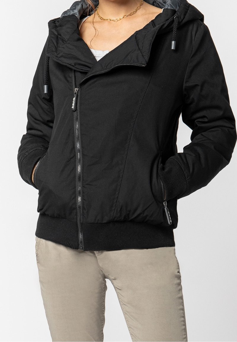 Femme portant une veste noire zippée avec des poignets et un ourlet côtelés, mains dans les poches, pantalon beige et un collier chaîne dorée visible au niveau du cou.