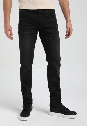 Homme portant un jean noir slim et des baskets noires à semelles blanches, debout devant un fond clair uni.