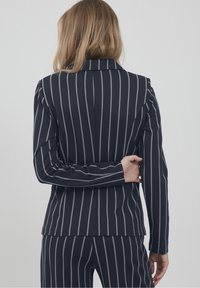 Femme aux cheveux blonds portant un costume bleu marine à rayures blanches verticales, debout avec un bras plié derrière le dos.