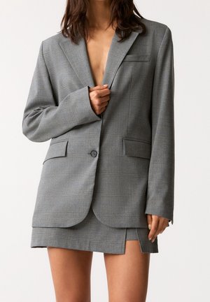Woman wearing a grey checked blazer and matching short skirt with a front slit, holding the blazer closed with one hand.