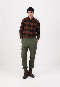 Brun og sort ternet skjorte med knapper, grønne cargo bukser med sidelommer og brune ankelstøvler, afsluttet med en sort strikket beanie.