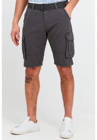 Man wearing dark gray cargo shorts with side pockets, white sneakers, and a tattoo on his left calf, standing on a neutral background.