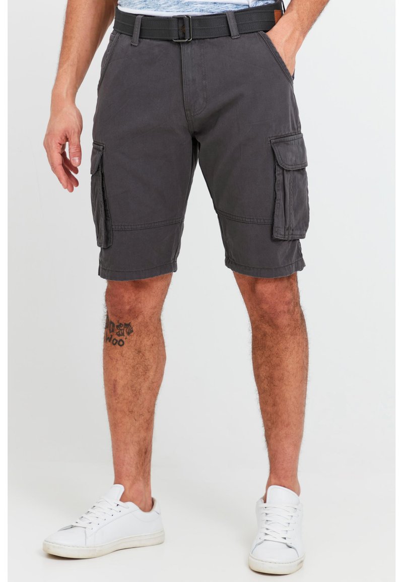 Man wearing dark gray cargo shorts with side pockets, white sneakers, and a tattoo on his left calf, standing on a neutral background.