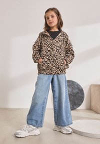 Jachetă cu fermoar cu imprimeu leopard dintr-un material moale, blugi largi albaștri și adidași argintii, stând pe un podea netedă cu forme geometrice.