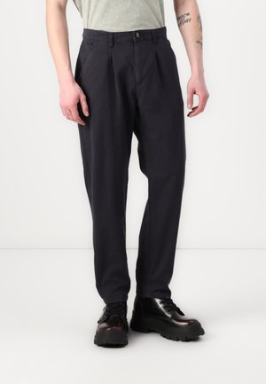 Pantalons chino bleu marine avec des plis, en coton, dotés d'une fermeture à bouton et de poches latérales, associés à des bottes à plateforme noires.