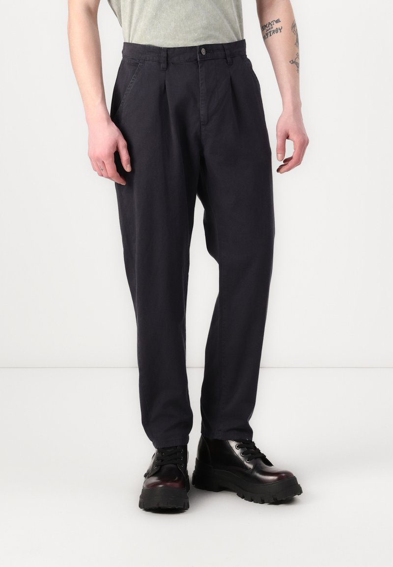 Pantalones chinos azul marino con pliegues, hechos de algodón, con cierre de botón y bolsillos laterales, combinados con botas de plataforma negras.