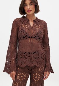 Blusa in cotone all'uncinetto marrone con intricati motivi floreali e geometrici, colletto e maniche lunghe a campana con dettagli aperti.