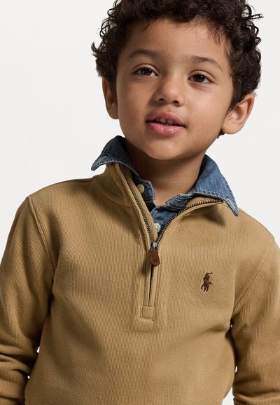 Tan ribbed pullover with a zip neck, layered over a denim shirt. Features a small brown logo on the chest and a textured fabric.