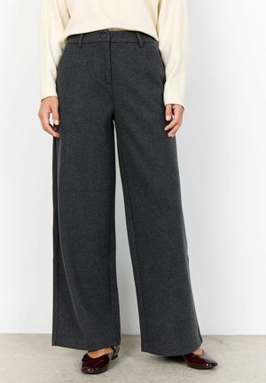 Vrouw in donkergrijze wijde broek, crèmekleurig langlijnig shirt en donkerbordeaux platte schoenen staat op een betonnen vloer.