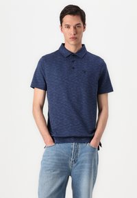 Camisa polo azul-marinho com um padrão texturizado, mangas curtas, gola com três botões e detalhe do logótipo, combinada com jeans azuis claros.