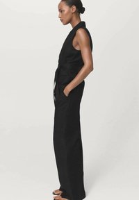 Donna in un jumpsuit nero senza maniche con cintura, in profilo con le mani nelle tasche, contro uno sfondo chiaro e neutro.