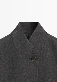 Szary płaszcz wełniany z kołnierzem o strukturalnym wykończeniu, z pojedynczym czarnym zapięciem na guzik i zarysowanym fasonem.