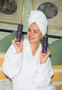 Due bottiglie cilindriche viola di prodotti per capelli tenute da una persona in accappatoio bianco e asciugamano. Sullo sfondo si vede una carta da parati verde decorativa e quadri incorniciati.