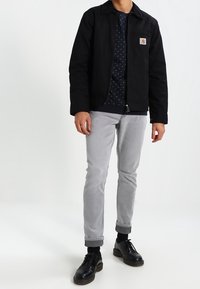 Giacca utility nera con zip, tasca sul petto, indossata sopra una T-shirt a fantasia blu navy, abbinata a pantaloni grigi chiari arrotolati e scarpe nere.