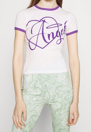 Kobieta nosząca biały krótki t-shirt z fioletowym obszyciem i napisem "Angel", w połączeniu z jasnymi zielonymi spodniami w marmurkowy wzór.