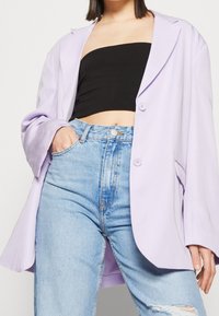 Světle fialový oversized blazer vyrobený z hladké tkaniny, s dvěma knoflíky, velkými klopami a kapsami, nošený přes černý crop top a modré džíny.
