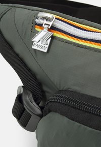Marsupio in nylon verde con dettagli delle zip multicolori, tirazip argentato e tracolla nera. Presenta hardware resistente e materiale testurizzato.
