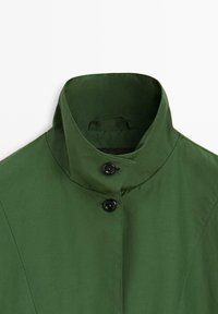 Groene jas met hoge kraag met twee zwarte knopen die de kraag sluiten op een witte achtergrond.