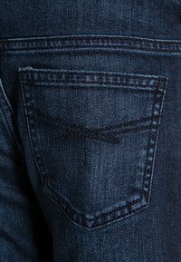 Tmavě modré džíny z denimu s kapsou na zadní straně a zakřivenou klopou, viditelným švem a texturovanou tkaninou.