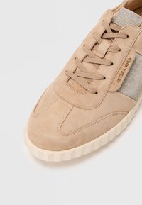 Beige suede sneaker with a textured elastic side panel and lace-up closure. Features a ridged rubber sole for grip.