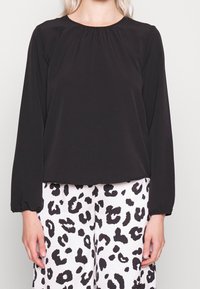 Schwarze Bluse mit langen Ärmeln, rundem Halsausschnitt und Raffung. Glatte Textur, Manschetten mit Elastik, kombiniert mit weißen Hosen im Leopardenmuster.