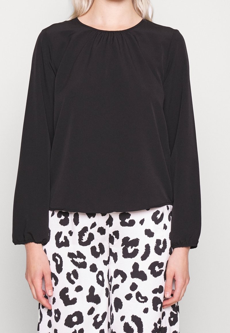 Schwarze Bluse mit langen Ärmeln, rundem Halsausschnitt und Raffung. Glatte Textur, Manschetten mit Elastik, kombiniert mit weißen Hosen im Leopardenmuster.