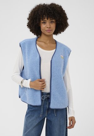 Frau mit lockigem Haar, die eine hellblaue Fleece-Weste über einem weißen Langarmshirt und hochgeschnittenen blauen Jeans mit einem gebundenen Gürtel trägt.