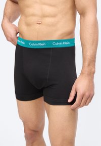 Férfi törzs fekete boxeralsót visel, amelynek türkizkék derékpántján a Calvin Klein logó látható, egyszínű világos háttér előtt áll.