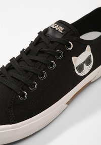 Musta kanvaslenkkari mustilla nauhoilla, valkoisella pohjalla ja valkoisella kissalla aurinkolaseissa sivuosassa.