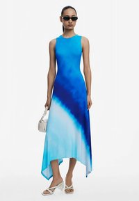 Niebieska i turkusowa sukienka tie-dye z asymetrycznym dołem, bez rękawów, wykonana z gładkiej tkaniny. Noszona z białymi sandałami i małą torebką.