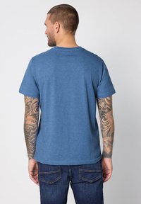 Blå t-shirt med rund hals i mjukt material, med korta ärmar och avslappnad passform, bärs med mörka jeans och visar arm tatueringar.