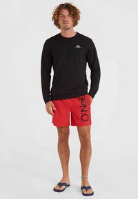 Zwarte sweatshirth met lange mouwen en een klein logo, gecombineerd met rode zwembroek met een zwart dessin. Draag met blauwe slippers.