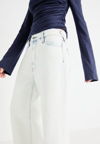 Jeans albi cu croi larg și spălare deschisă, având talie înaltă, buzunare frontale și cusături vizibile. Asortați cu un top bleumarin cu mânecă lungă.