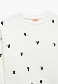 Witte sweatshirt met zwarte hartpatronen, ronde halslijn en zachte textuur. Label zichtbaar met "baby" merk.