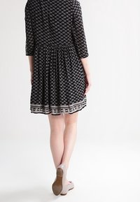 Vestido negro con un patrón geométrico blanco, mangas 3/4, cintura ceñida, falda acampanada y detalle de encaje en el dobladillo. Llevado con zapatos de color claro.